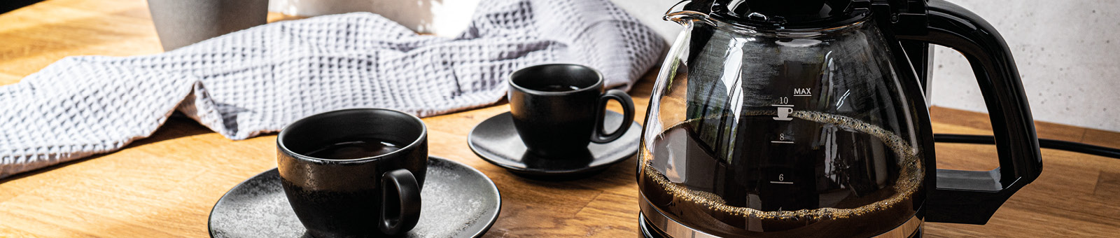 Das Bild zeigt zwei Kaffeetassen, eine Kaffeekanne und im Hintergrund eine Kaffeemaschine