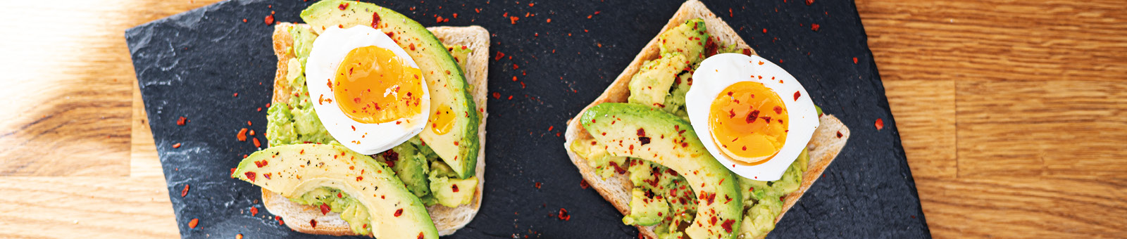 Das Bild zeigt aus der Vogelperspektive zwei Scheiben Toast belegt mit Avocado und Ei auf einer Schieferplatte.