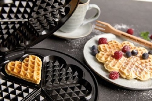Das Bild zeigt das Waffeleisen Cloer 1898 mit einem Waffelherz, daneben steht ein Teller mit einer Herzwaffel, dekoriert mit Beeren und daneben eine graue Tasse.
