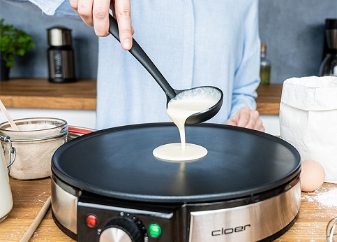 Das Bild zeigt den Crêpes Maker Cloer-6630 und eine Hand die mit einer Kelle Teig auf die Backfläche gibt.