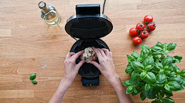 Das Bild zeigt aus der Vogelperspektive zwei Hände über einer Holzarbeitsplatte, die eine Teigkugel in ein Waffeleisen gibt, daneben Tomaten, Basilikum und eine Flasche Öl. 