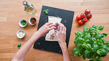 Das Bild zeigt aus der Vogelperspektive zwei Hände über einer Holzarbeitsplatte,  die Schinkenwürfel auf ein ausgerolltes Teigstück gibt, daneben Tomaten, Basilikum, Töpfchen mit Gewürzen und eine Flasche Öl. 