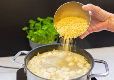 Das Bild zeigt wie in einen Suppentopf mit Suppe, Suppennudeln gegeben werden.